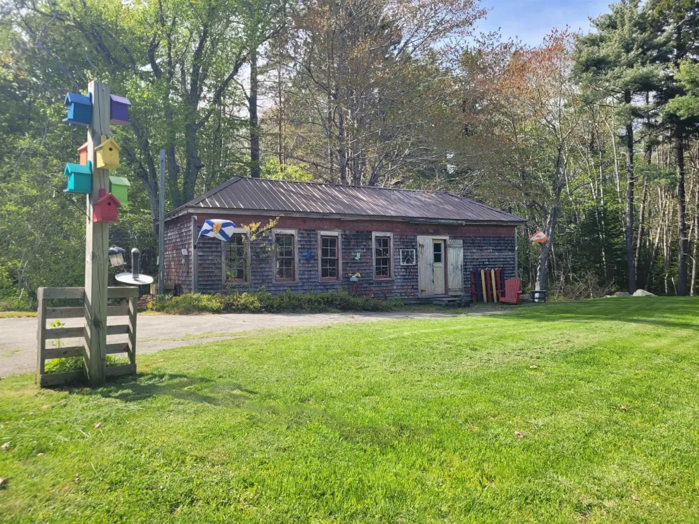 Rustic building with a metal roof and weathered wood siding, surrounded by green grass and trees. A colorful birdhouse post stands nearby.