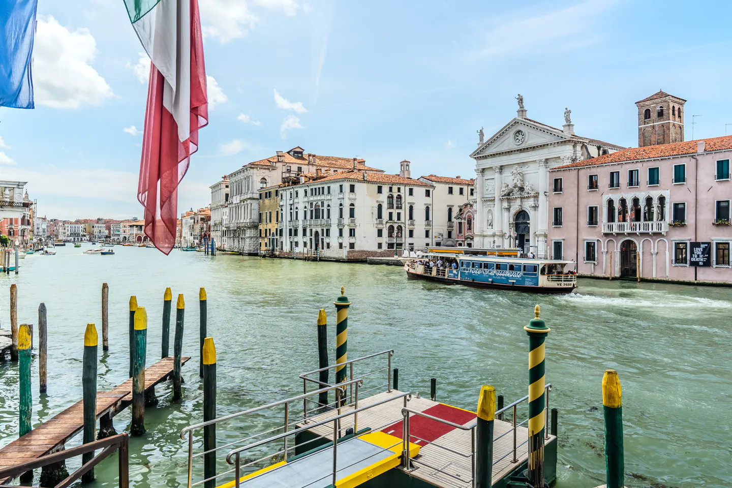 A view of a Venice canal with a water bus, historic buildings, and striped mooring poles under a bright blue sky.