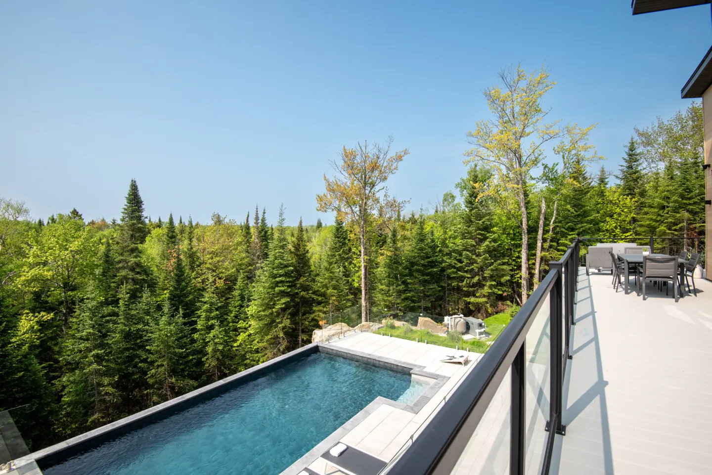 A modern home's deck overlooks a forest and an infinity pool. The deck has a glass railing and outdoor furniture.
