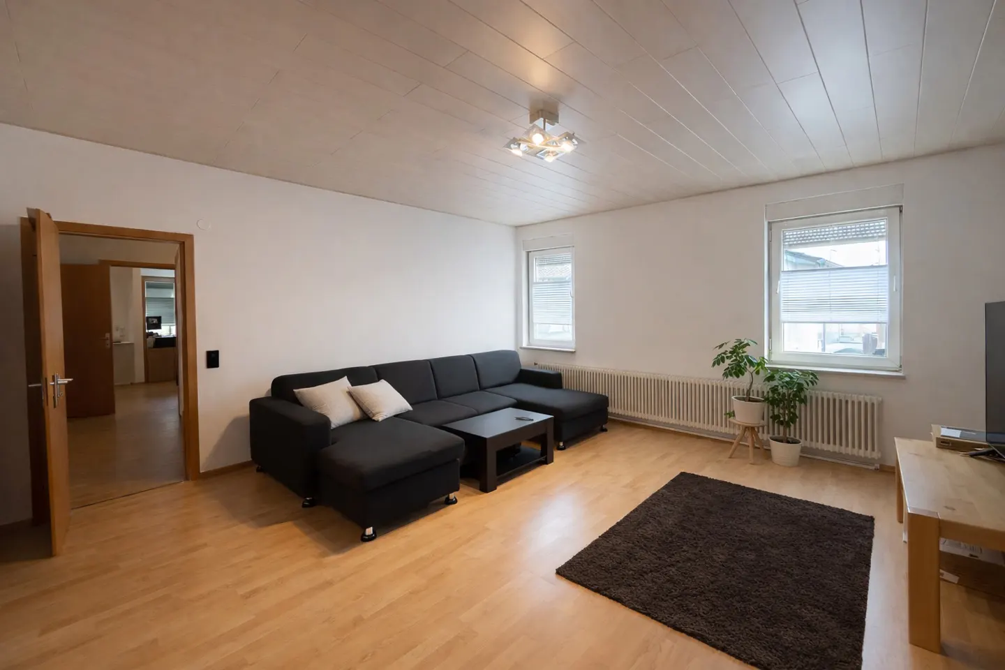 Living room with wood floors, white walls, and a black sectional sofa with light pillows. A dark rug sits in front of a TV stand.