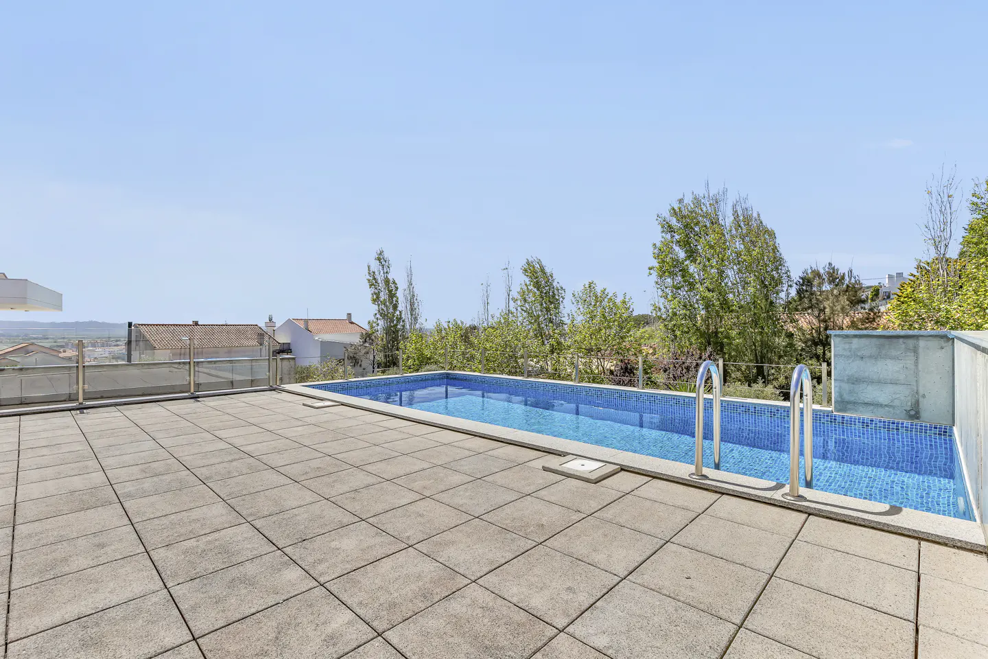 Outdoor pool with blue tiles and metal ladder, surrounded by a stone patio. Trees and houses are visible in the background under a clear blue sky.