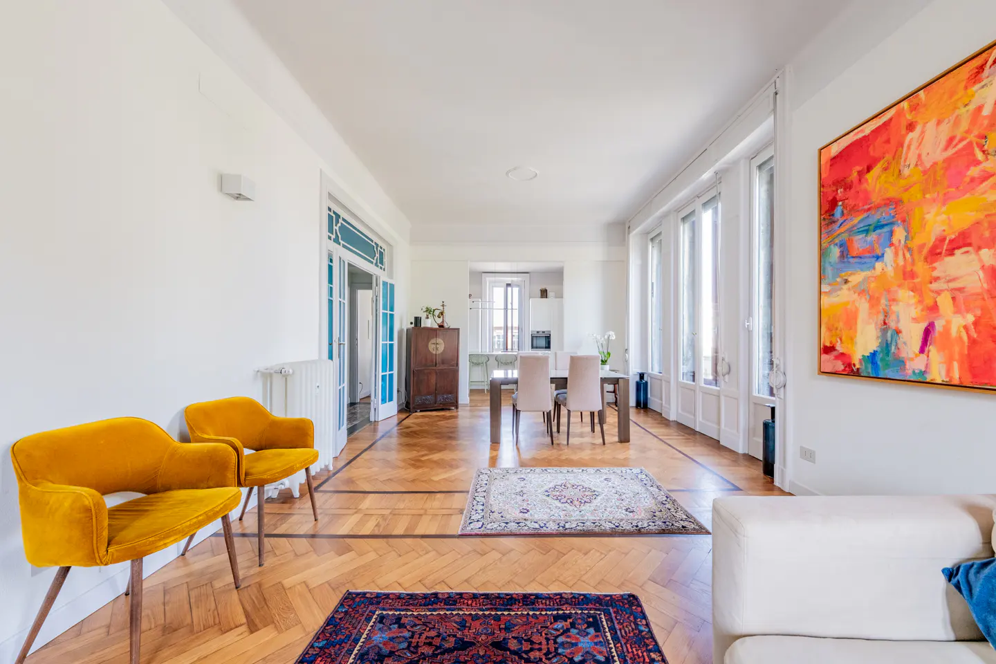 Bright, airy living room with herringbone wood floors, yellow chairs, and a colorful abstract painting. Dining table and kitchen visible.