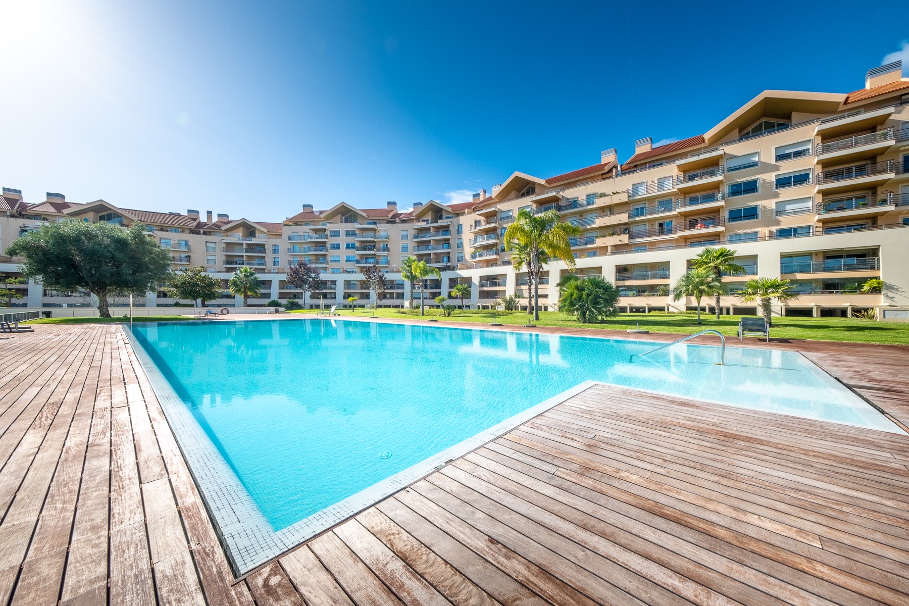 Large swimming pool with wooden deck, surrounded by a multi-story apartment complex, palm trees, and a clear blue sky. [AI-generated text]
