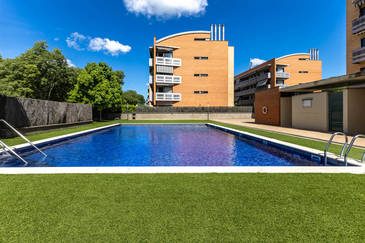 A rectangular swimming pool with blue water, surrounded by green grass and apartment buildings under a blue sky.