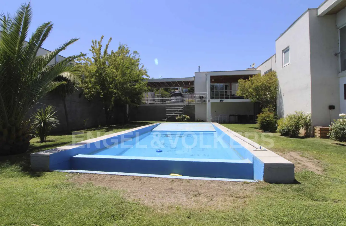 A backyard with a blue swimming pool, green grass, trees, and a white two-story house on a sunny day.