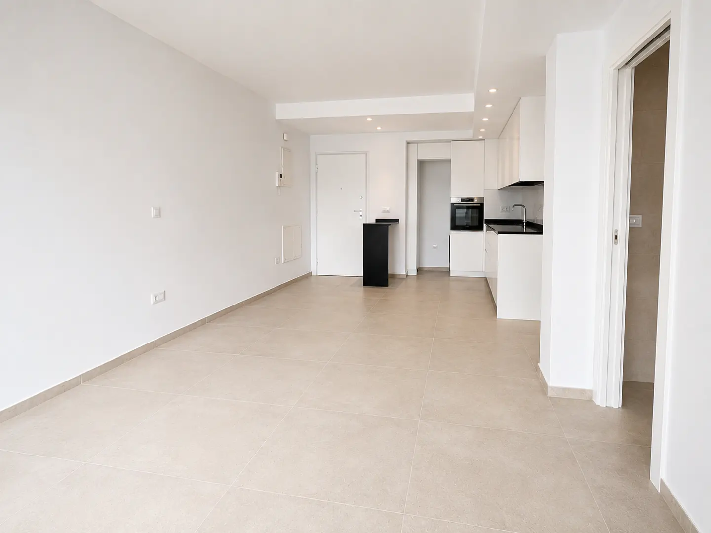 Bright, empty apartment with white walls, beige tile floor, and a modern white kitchen with black countertops. A black table stands near the entrance door.
