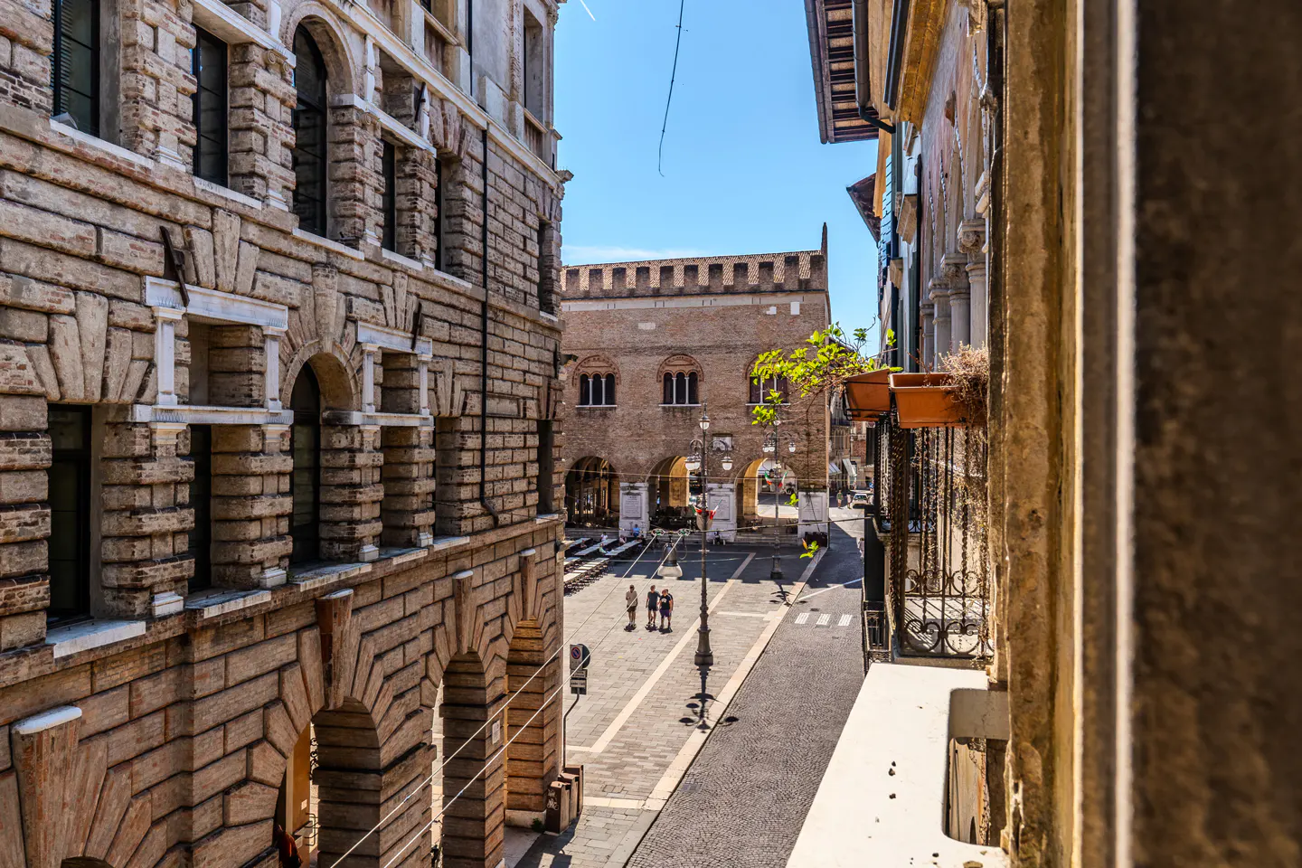 View from a balcony overlooking a sunny Italian-style cobblestone square framed by historic brick buildings with arches.