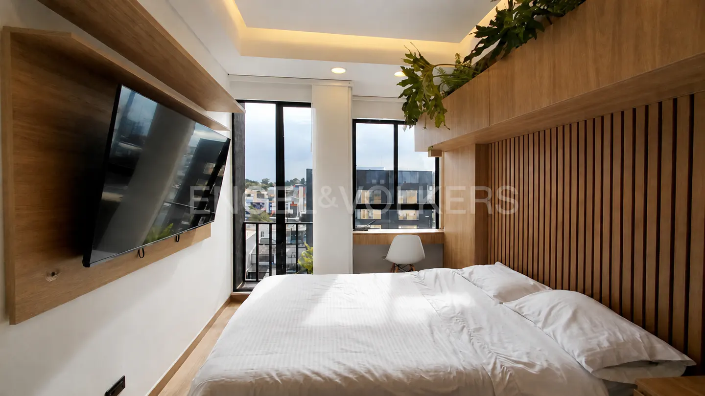 Modern bedroom with a white bed, wood-paneled wall, wall-mounted TV, and a small desk by the window with city views.