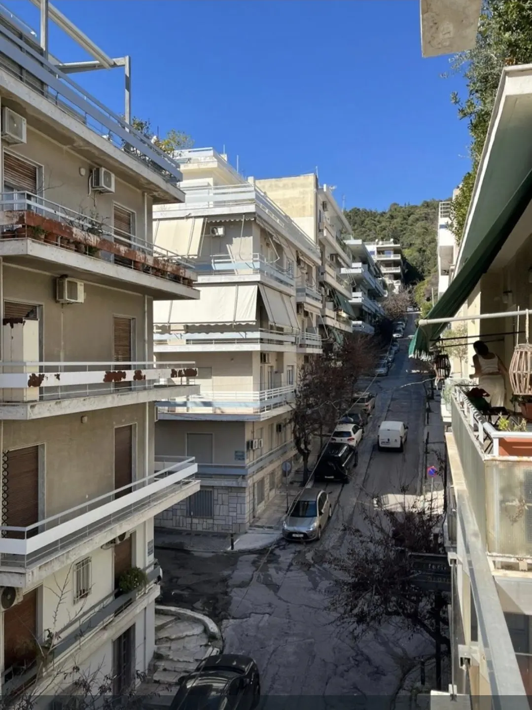 Street view of a neighborhood with beige apartment buildings, parked cars, and a blue sky.