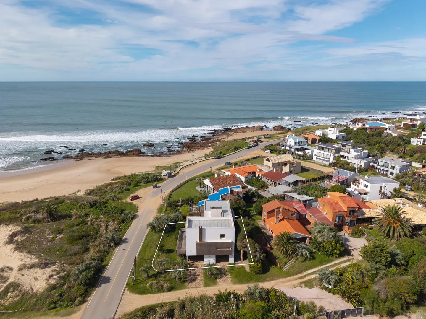 Aerial view of a modern white and brown house near a sandy beach and blue ocean under a partly cloudy sky.