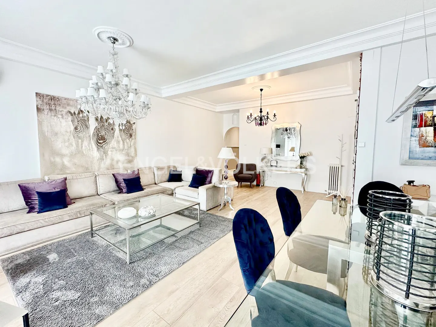 Elegant living room with a large beige sectional, glass coffee table, crystal chandelier, and blue velvet dining chairs.