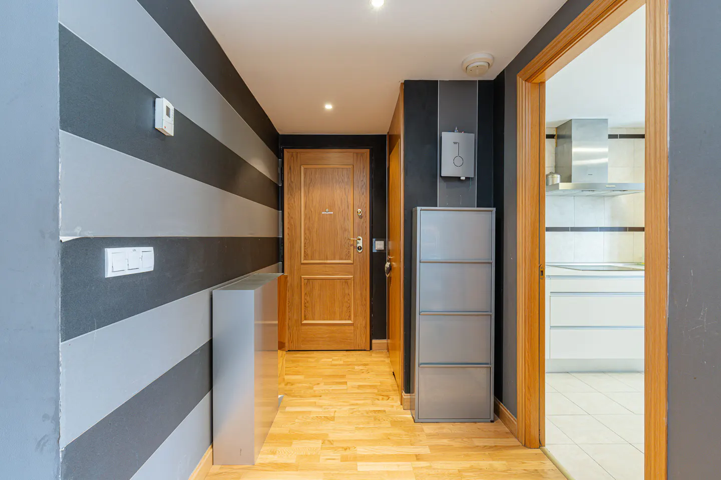 Hallway with gray and silver striped walls, a wooden door, and a glimpse of a white kitchen through an open doorway.