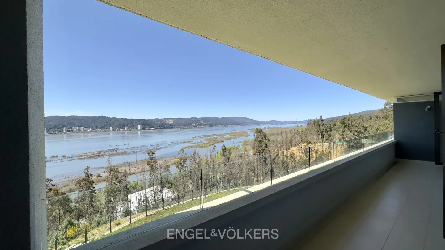 A modern balcony with a glass railing overlooks a wide river, lush trees, and distant hills under a clear blue sky.