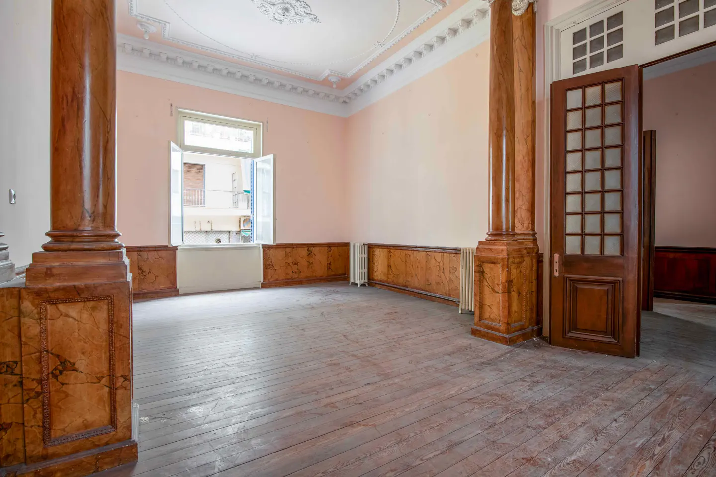 Empty room with pink walls, wood floors, and tall wood columns. A window is open, and a door leads to another room.