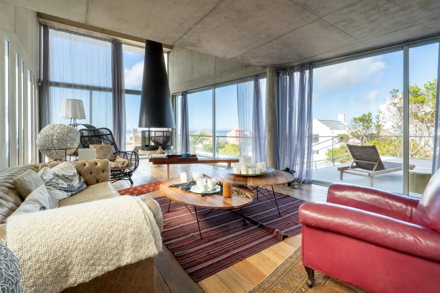 Bright living room with floor-to-ceiling windows, a black fireplace, and a red leather chair. A wooden coffee table sits on a striped rug.
