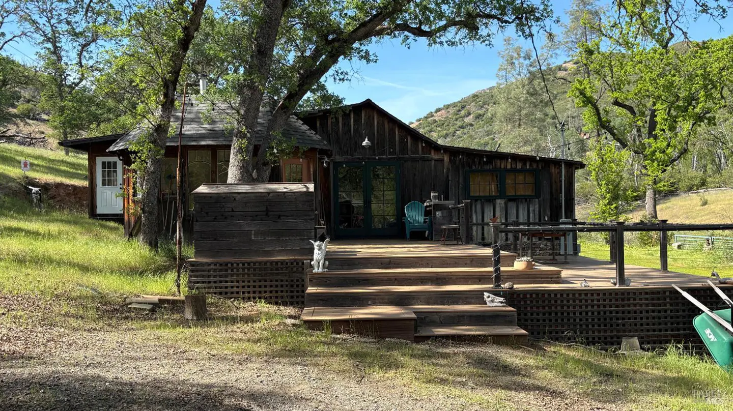 Rustic wooden cabin with a deck and stairs, nestled among trees and a grassy yard. A mountain is visible in the background.