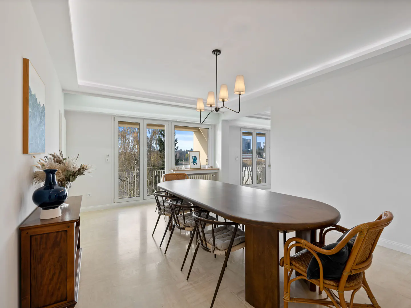 Bright dining room with a long wooden table, rattan chairs, modern chandelier, and large windows overlooking trees.