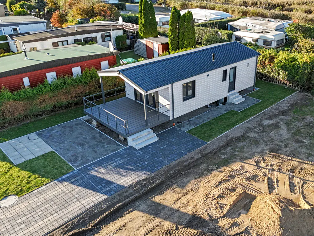 Aerial view of a modern, white mobile home with a blue roof and gray deck, surrounded by green grass and a paved driveway.