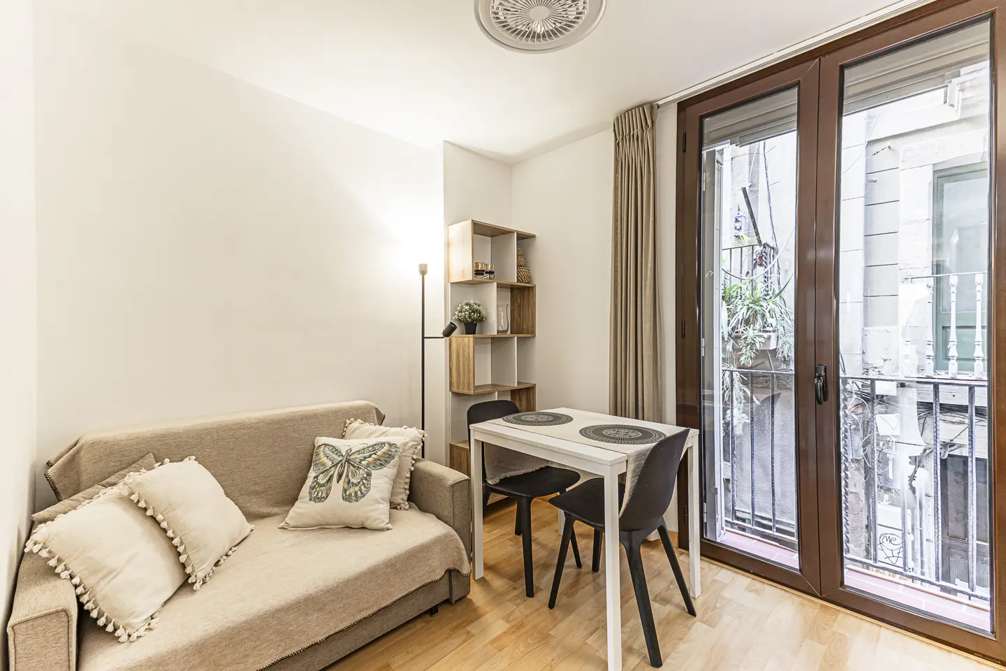 Living room with a beige sofa, white pillows, a white table with two black chairs, and a balcony with brown doors.