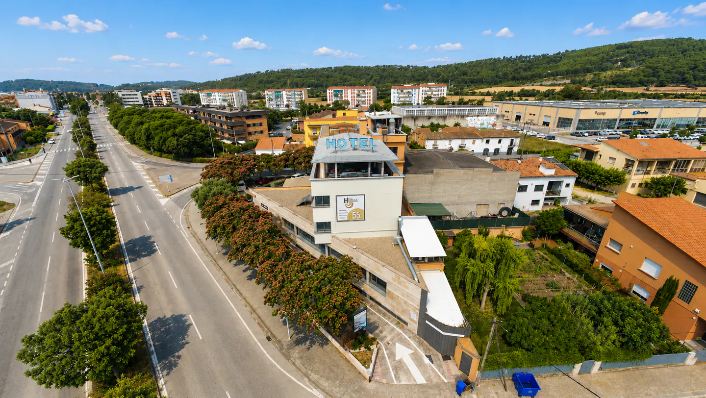 Aerial view of a hotel building with a blue sign, situated next to a road, surrounded by trees and residential buildings.