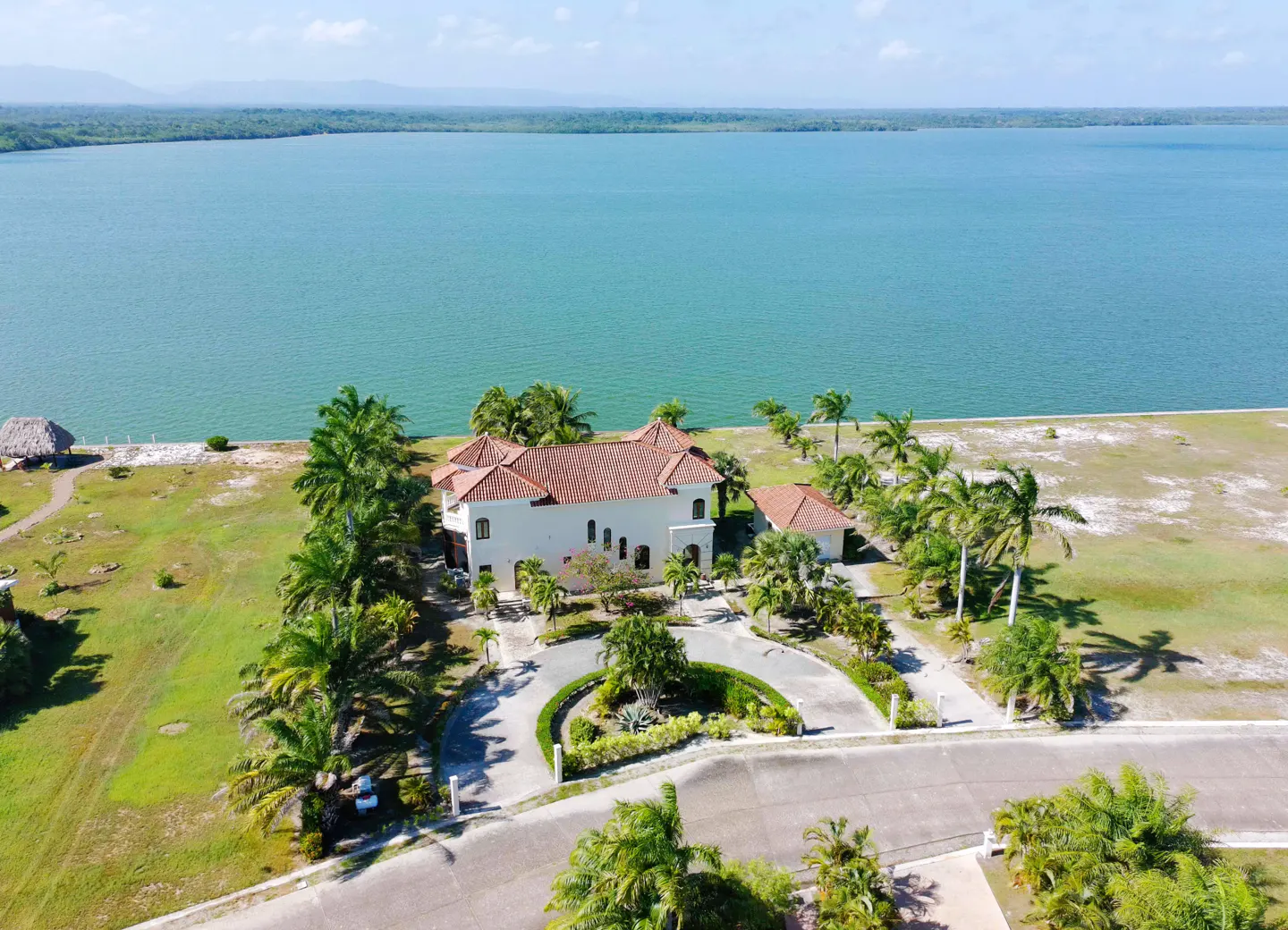 Aerial view of a white house with a red tile roof, palm trees, and a circular driveway near a large body of water.