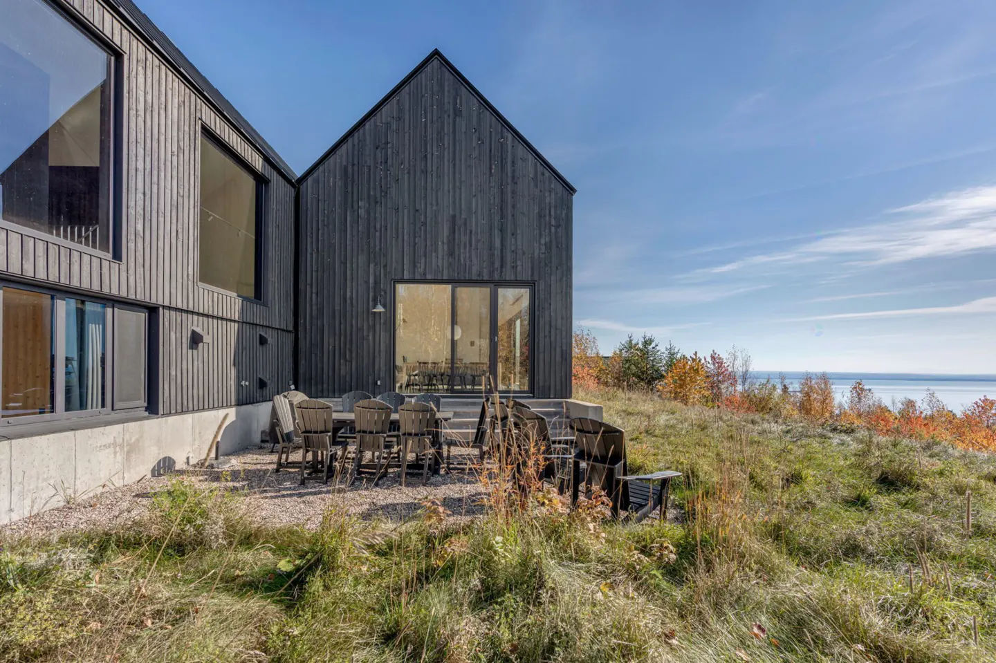 Modern black house with a patio, table, and chairs overlooking a grassy hill and a body of water under a blue sky.