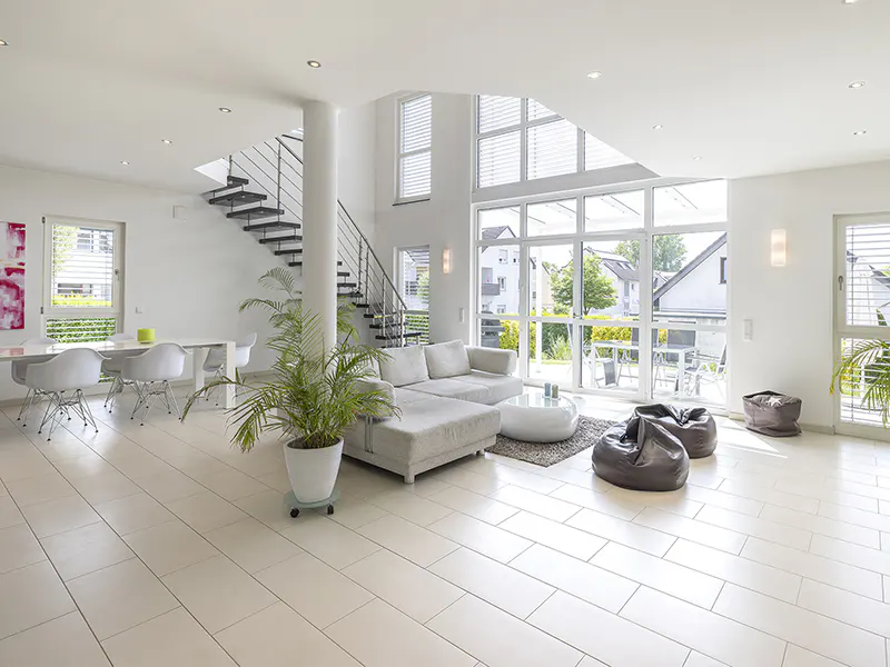 Bright, modern living room with white tile floors, a gray sofa, beanbag chairs, and a staircase. Large windows offer a view of the outdoors.