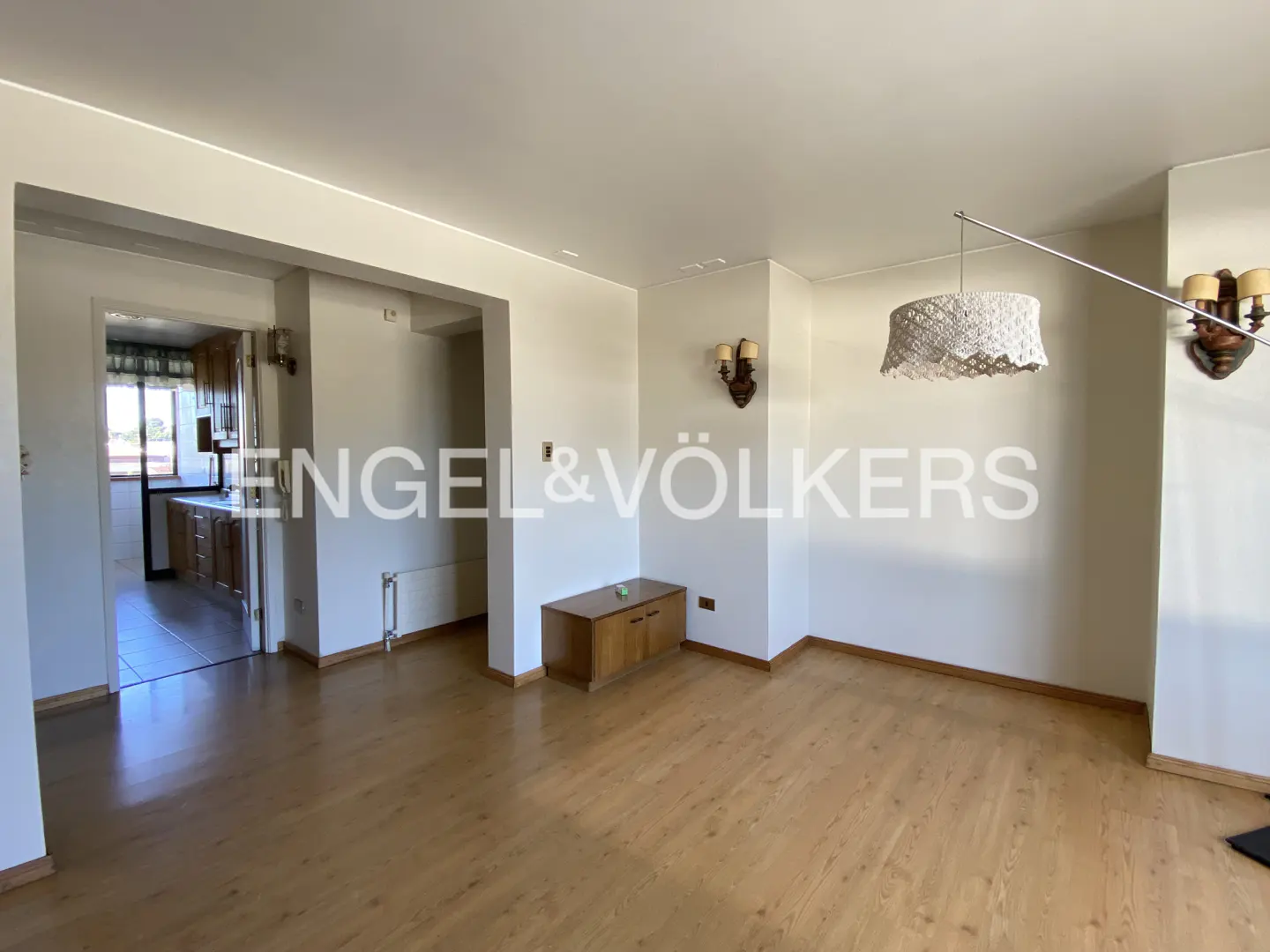 A bright living room with wood floors, white walls, and a view into the kitchen. A wooden cabinet sits against the wall.