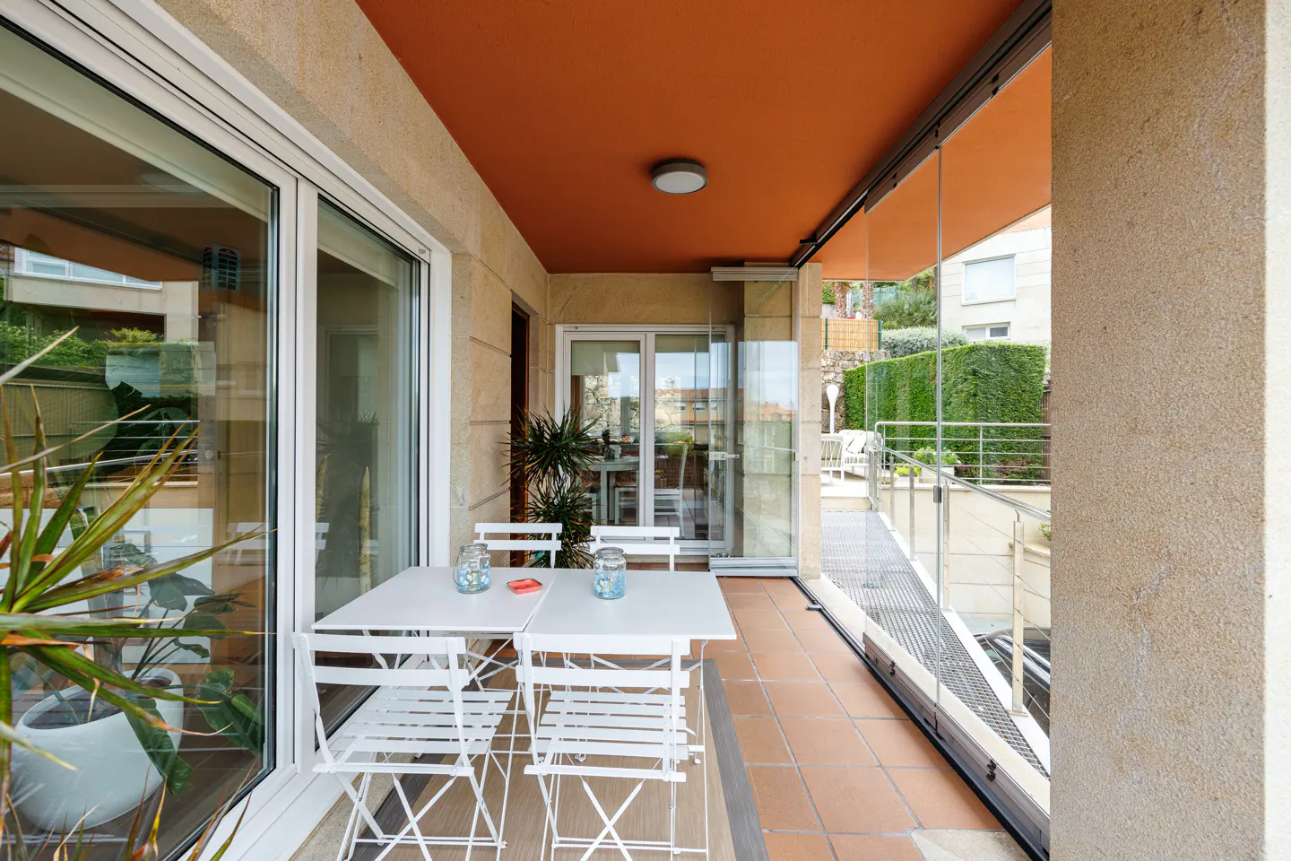 A sunlit balcony with white metal furniture, glass walls, and a terracotta-tiled floor. A view of greenery is visible beyond the balcony.