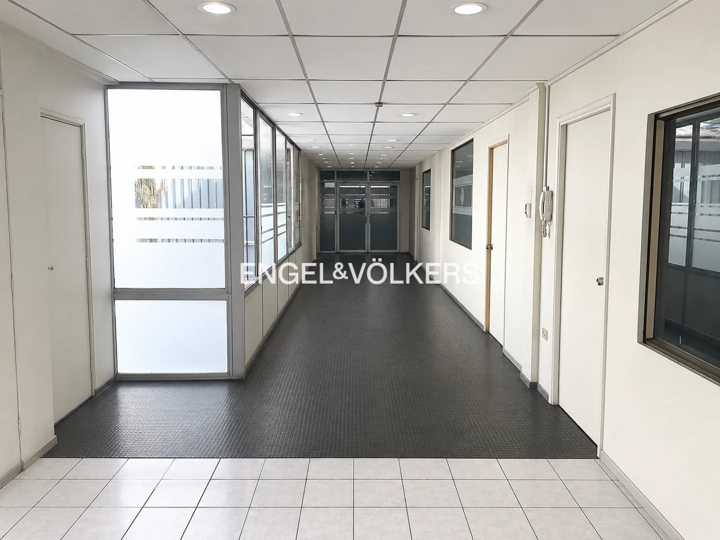 A long, bright hallway with white walls, doors, and a gray floor. Windows line the left side, and the Engel & Völkers logo is visible.