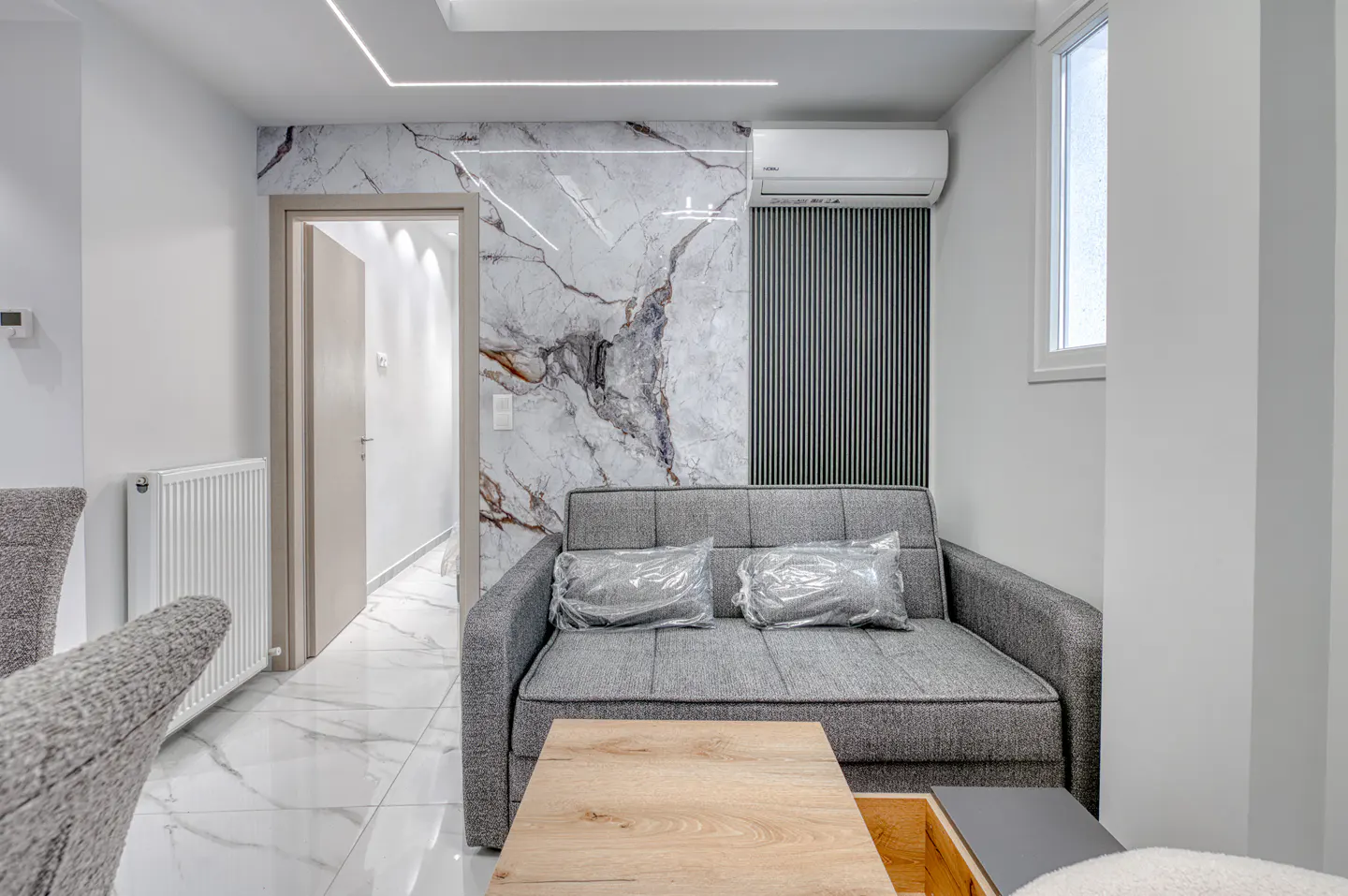 Living room with gray sofa, wood table, marble accent wall, and white walls. Pillows are wrapped in plastic.