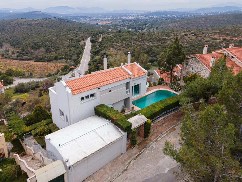 Aerial view of a modern white house with a red tile roof and a blue swimming pool, surrounded by green trees and hills.