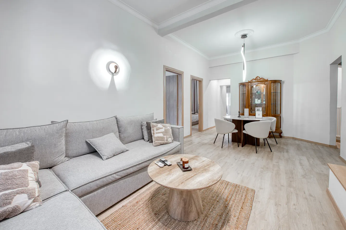 Bright living room with gray sectional sofa, round wood table, and dining area with antique cabinet and white chairs.