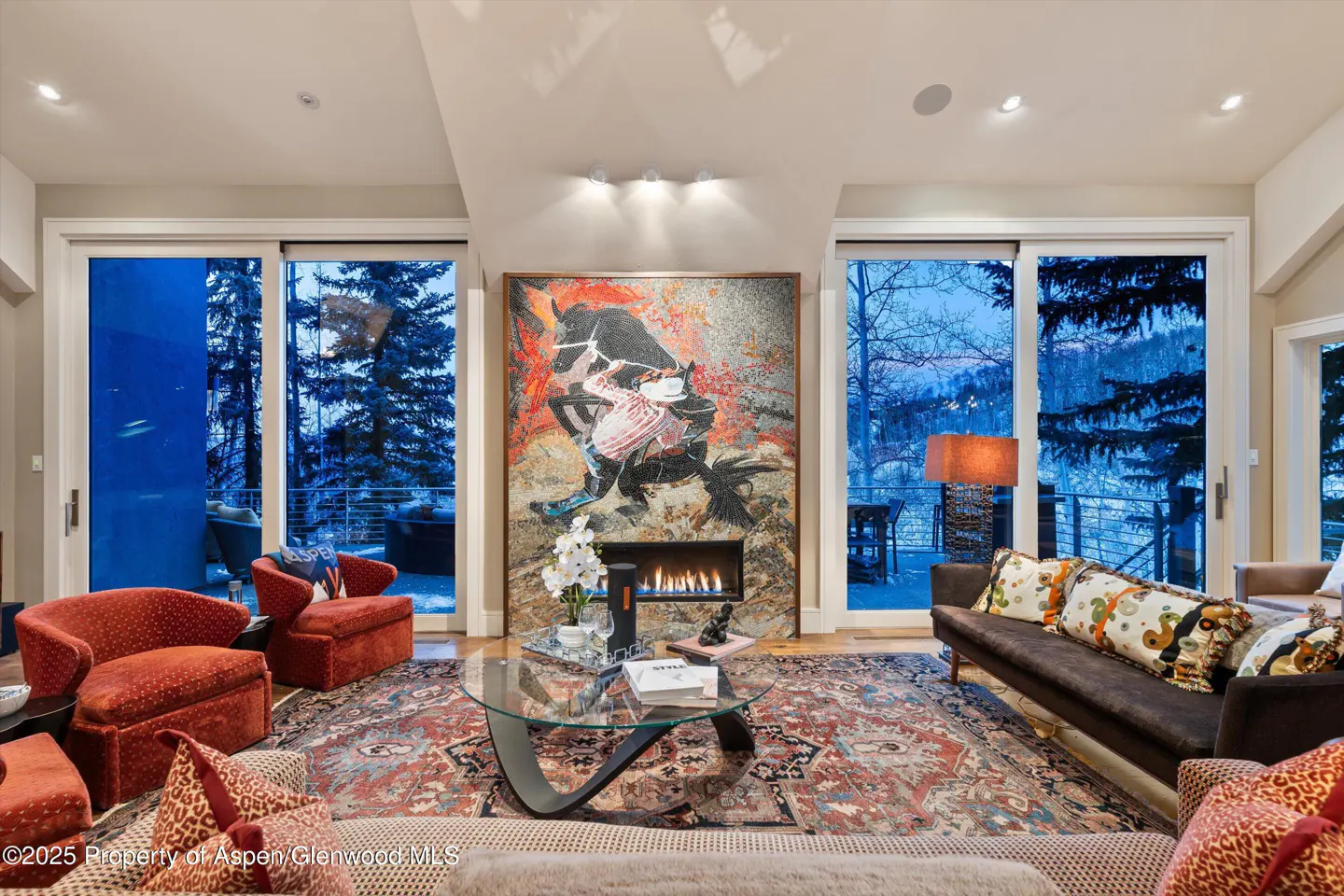 Living room with a fireplace, abstract art, and sliding glass doors to a snowy outdoor view. Red chairs and patterned rug add warmth.