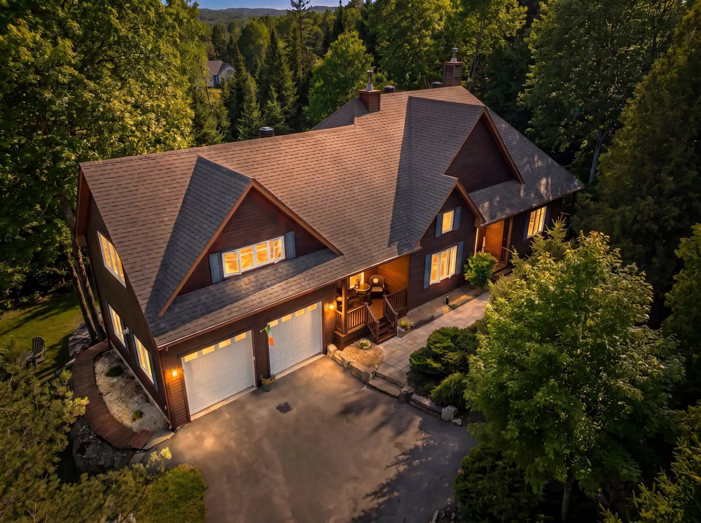 Aerial view of a brown house with a gray roof, white garage doors, and blue shutters, surrounded by green trees.