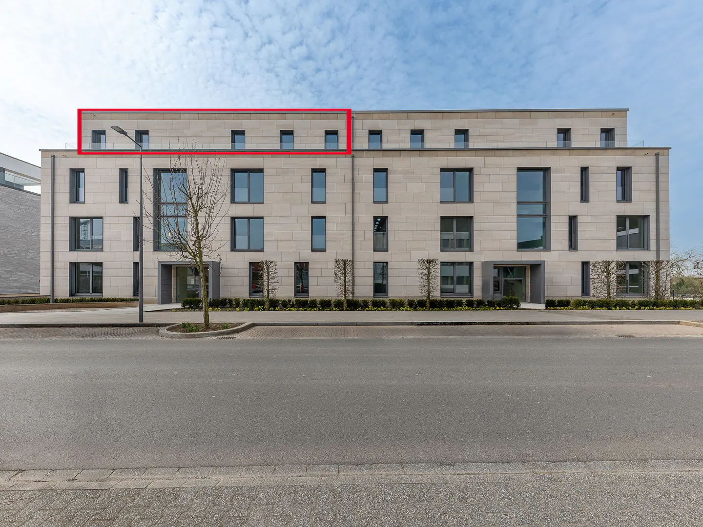 Modern, four-story apartment building with light stone facade and dark-framed windows, viewed from across a gray asphalt street.