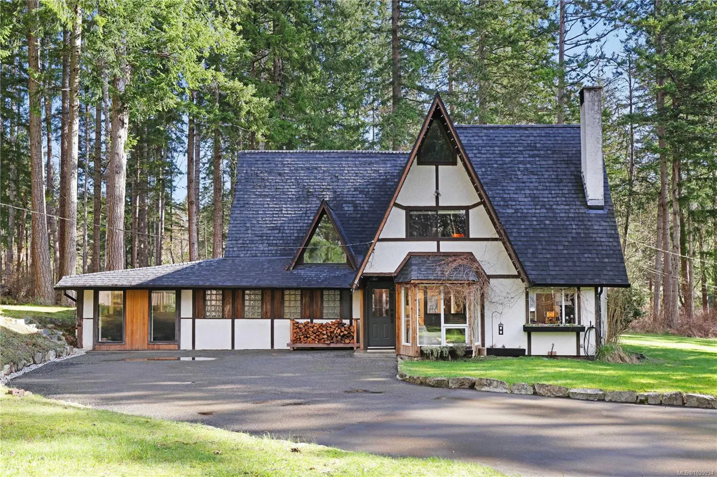 A-frame house with a dark roof, white walls, and wood trim, surrounded by tall trees and green grass.