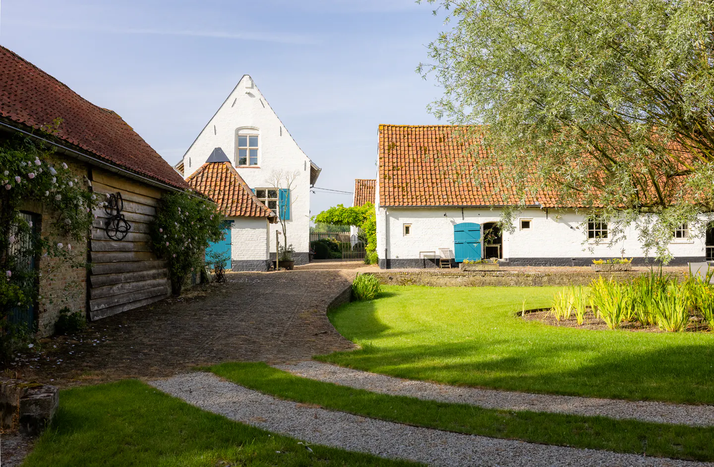 White farm buildings with orange tile roofs, blue doors, and a lush green lawn with a gravel path under a sunny sky.