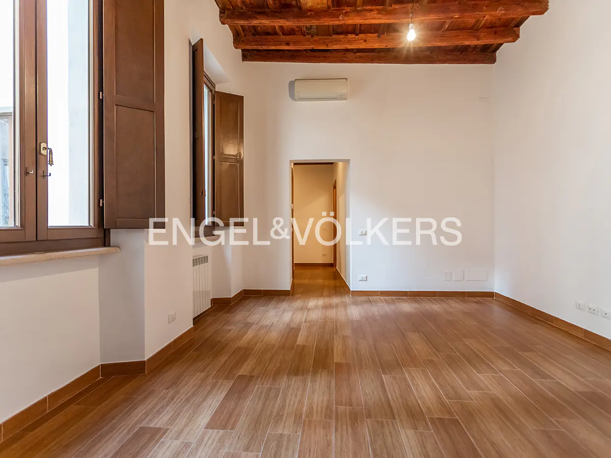 An empty room with wood floors, white walls, and brown window shutters. A doorway leads to another room. The ceiling has exposed wood beams.
