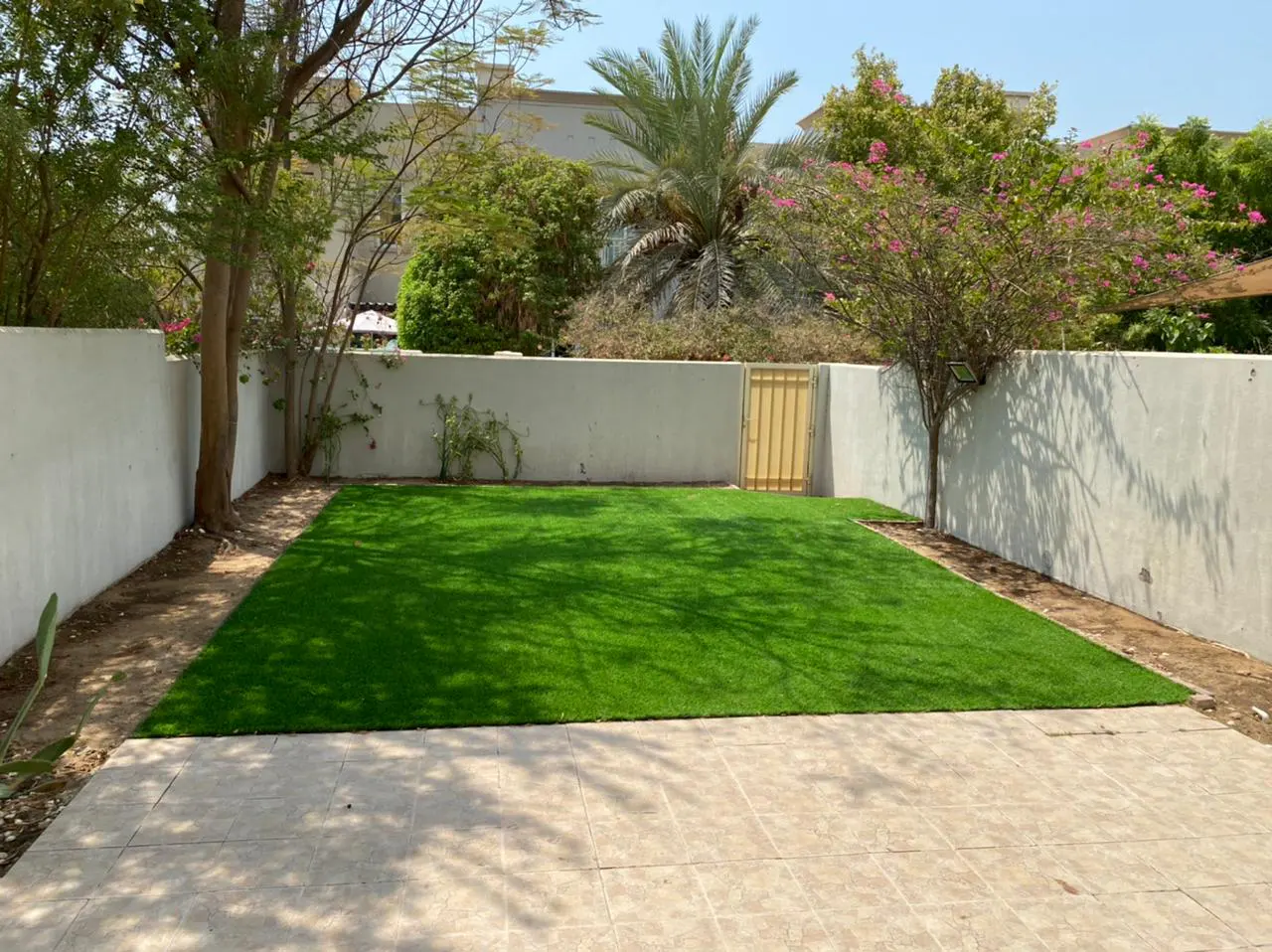A backyard with green grass, a beige tiled patio, and a white wall with a yellow gate. Trees and plants surround the yard.
