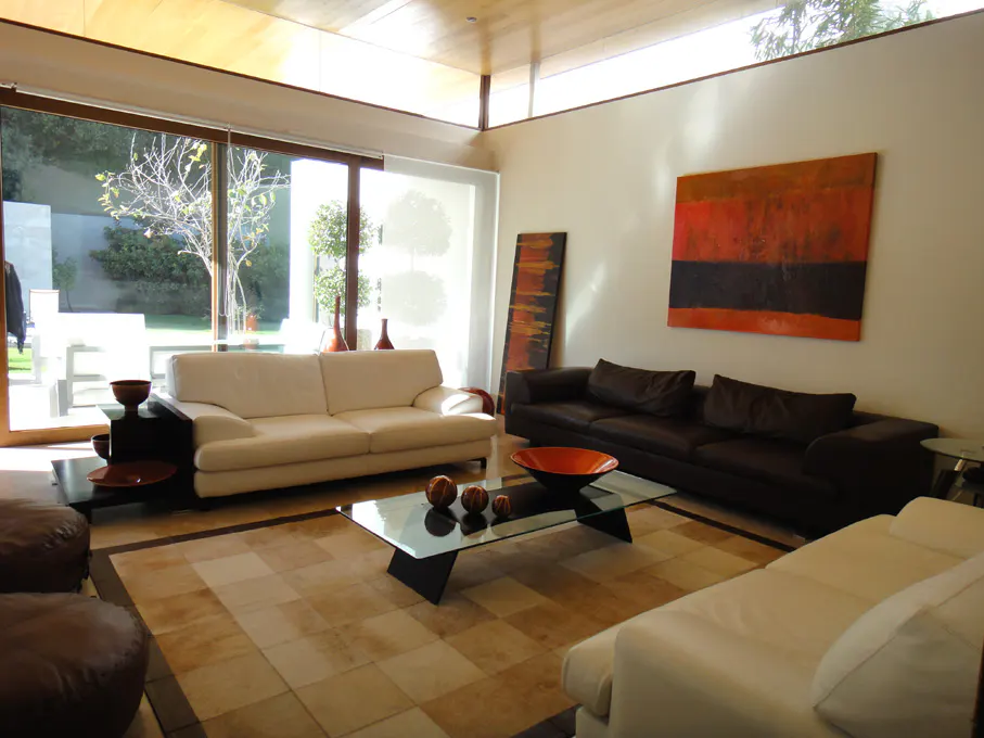 Bright living room with a white sofa, a brown sofa, and a glass coffee table on a checkered rug. Large windows overlook a green garden.