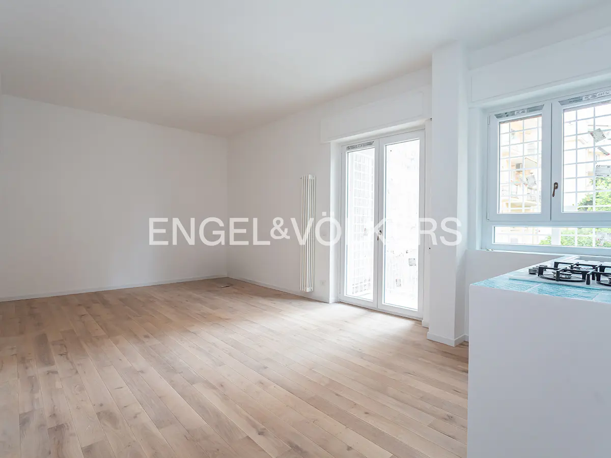 Bright, empty room with light wood floors, white walls, and a white radiator. Glass doors lead to a balcony. A kitchen area is visible with a blue tiled counter.