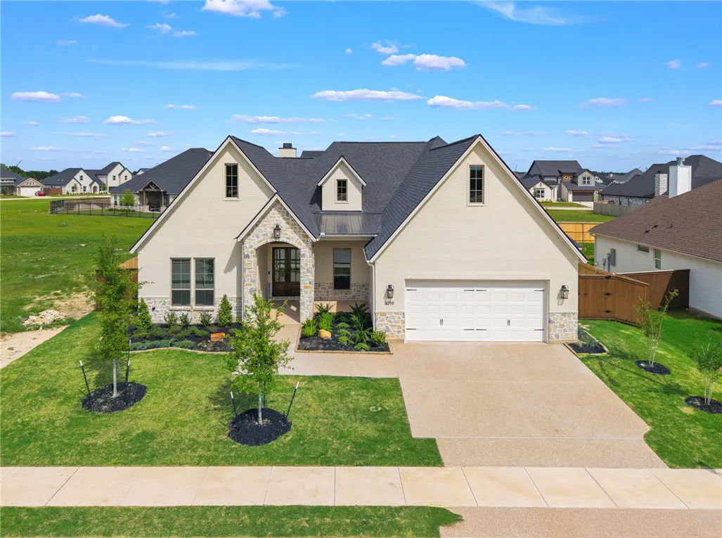 Beige single-story house with a dark gray roof, stone accents, and a two-car garage on a green lawn under a blue sky.