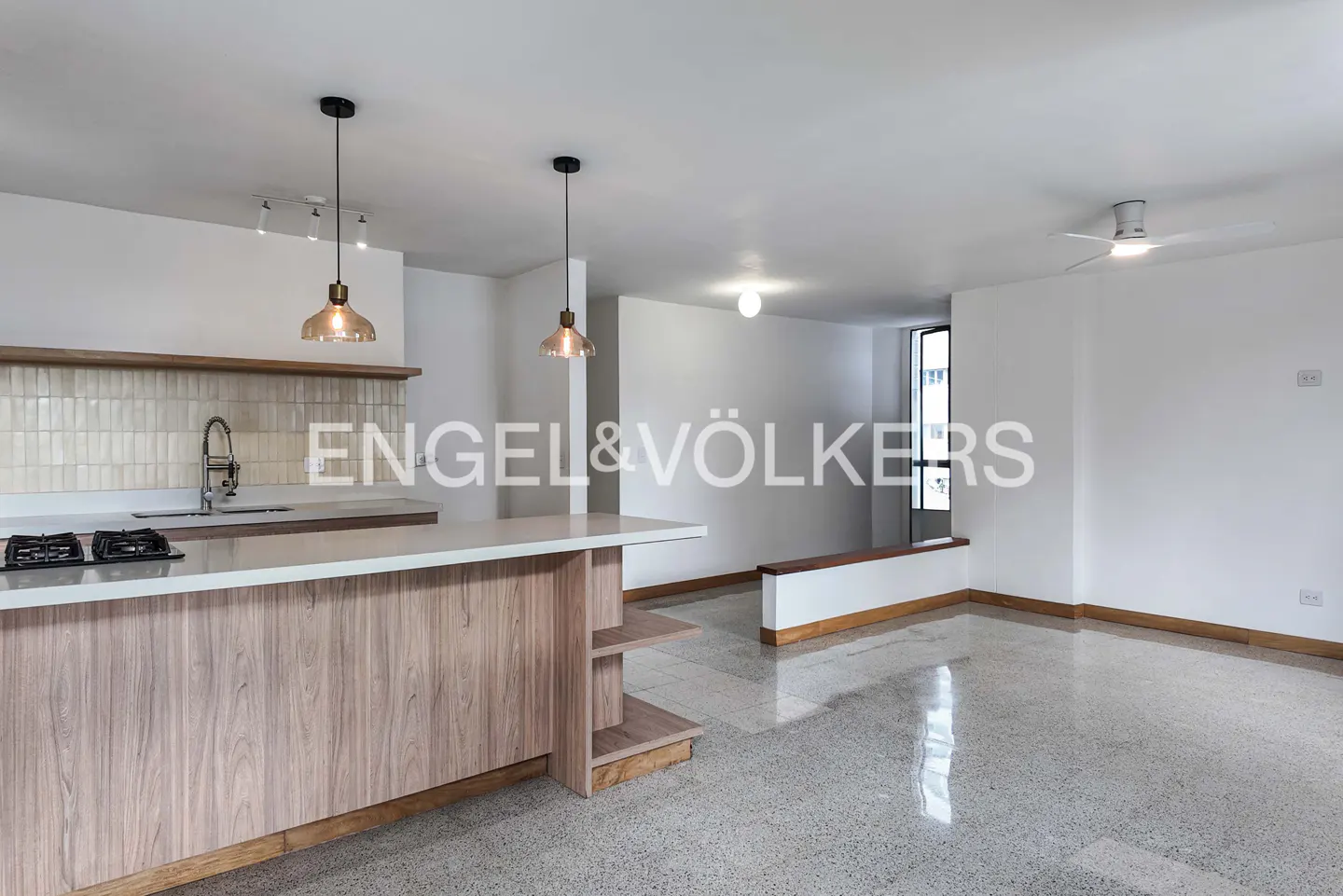 Open-concept kitchen and living room with wood cabinets, white walls, and terrazzo floors. Pendant lights hang over the kitchen island.