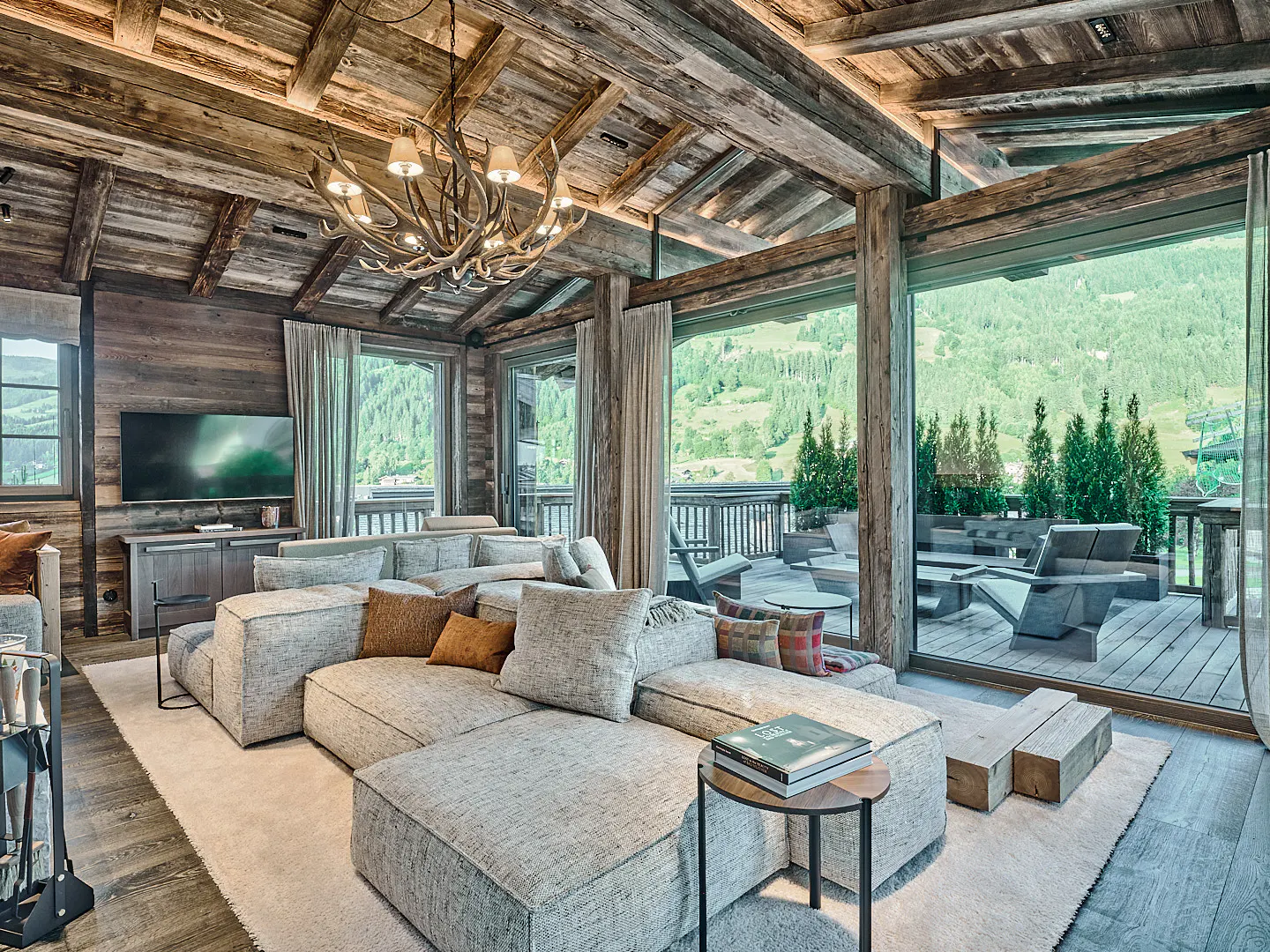 Rustic living room with wood walls, beams, and antler chandelier. Large sectional sofa faces a deck with mountain views.