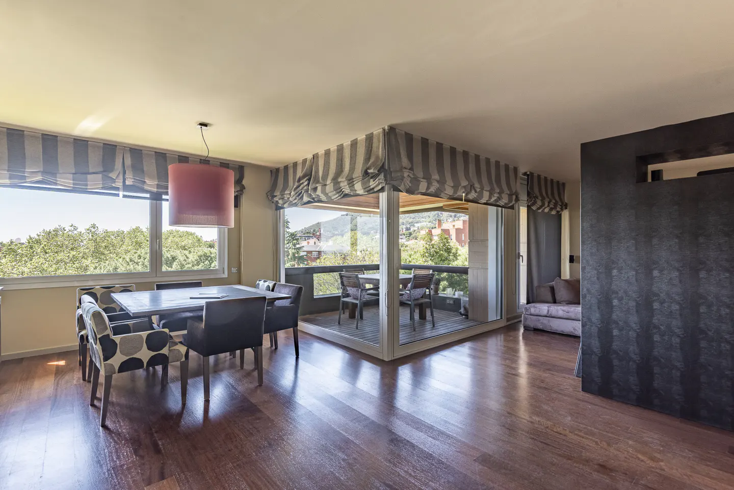 Bright dining room with wood floors, a table with chairs, and a balcony with outdoor seating overlooking a green landscape.
