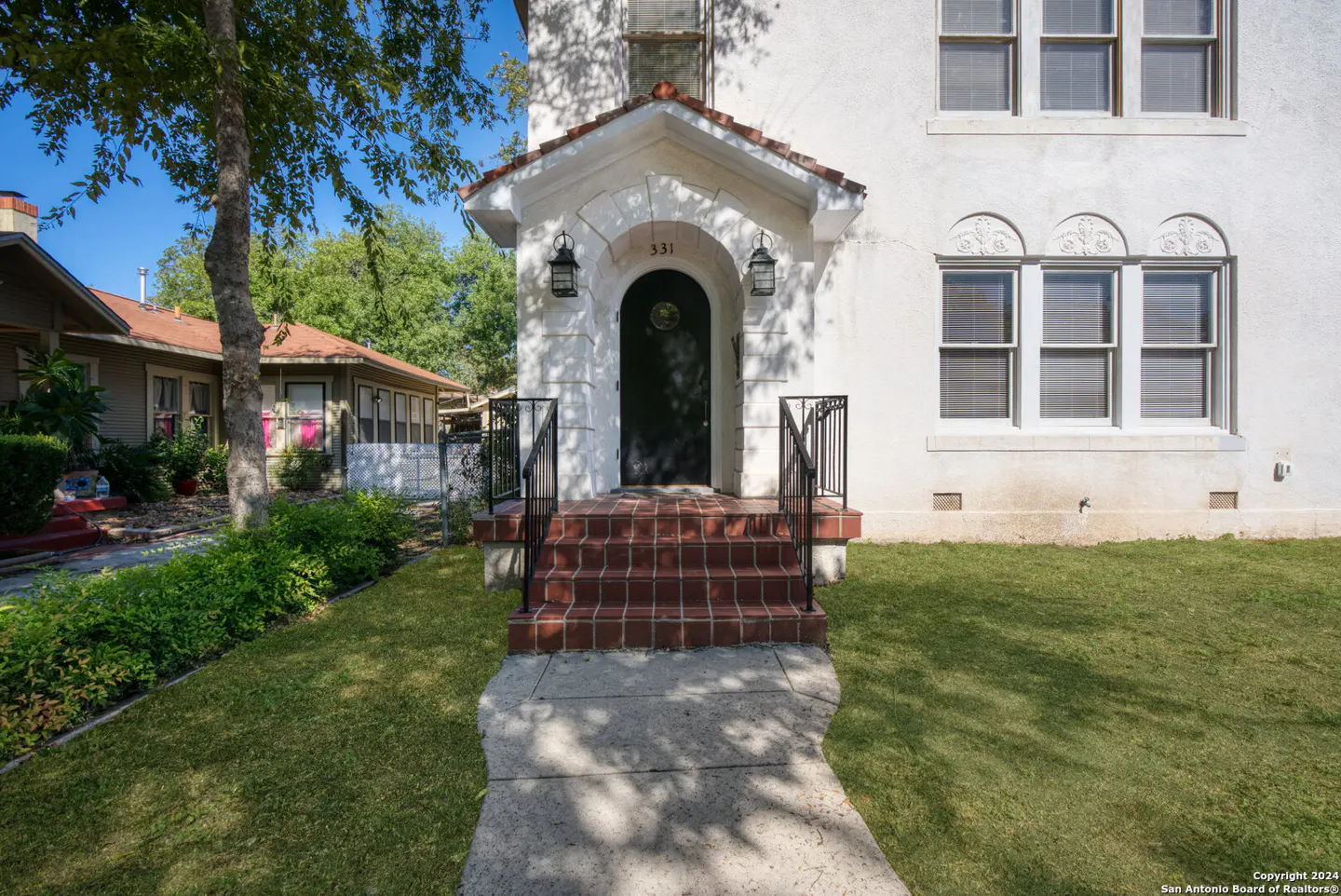 White stucco house exterior with a black arched front door, brick steps, and a concrete walkway on a sunny day.