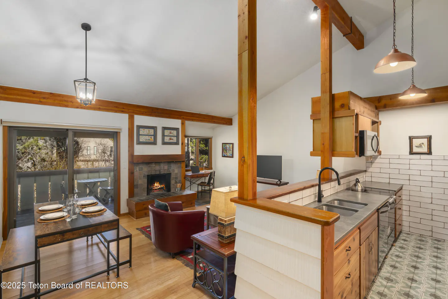 Open-concept living space with wood beams, fireplace, and kitchen. Dining table set for four near sliding glass doors to a balcony.