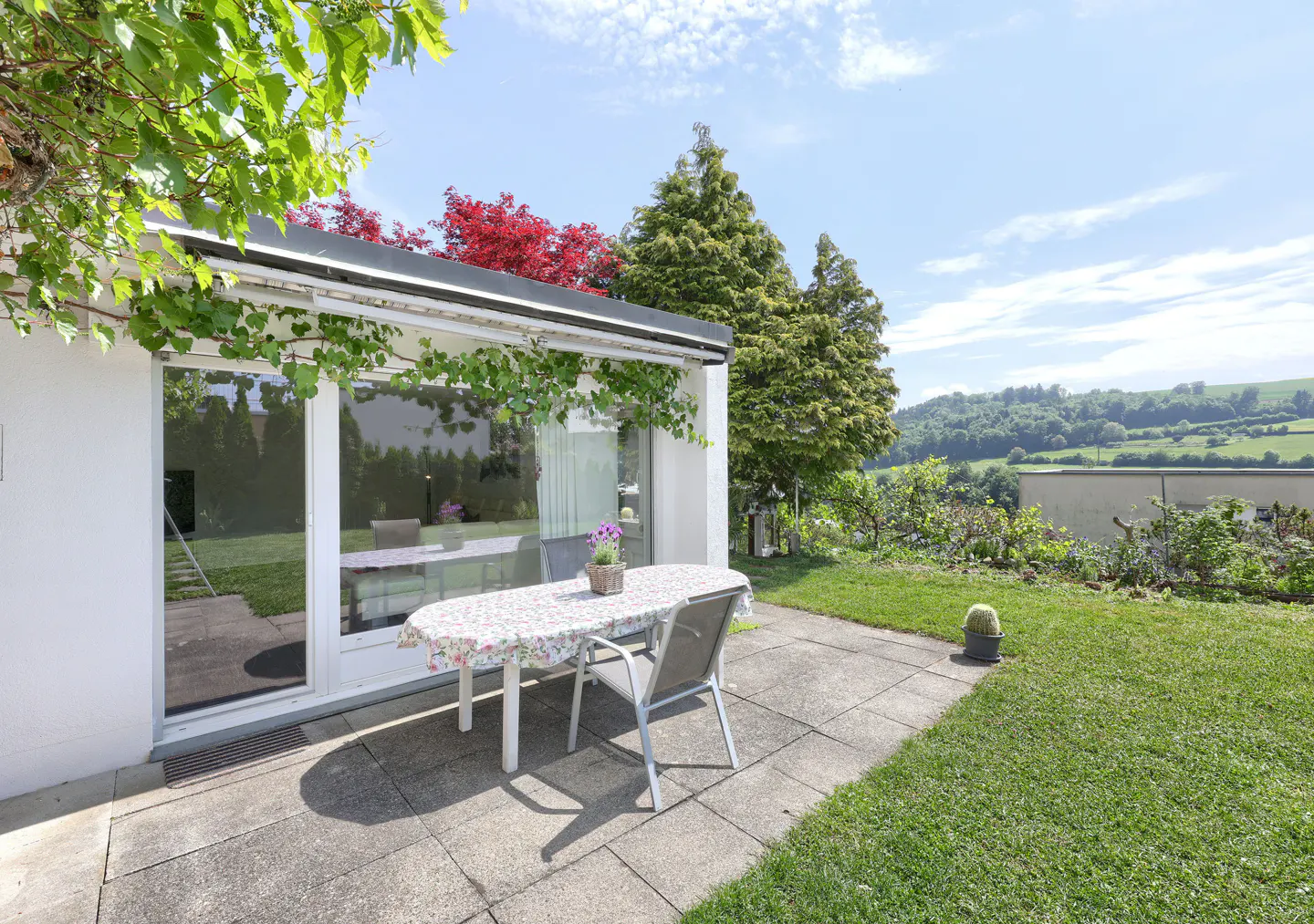 Patio with a floral-covered table and chairs outside a white house, overlooking a green lawn and rolling hills.