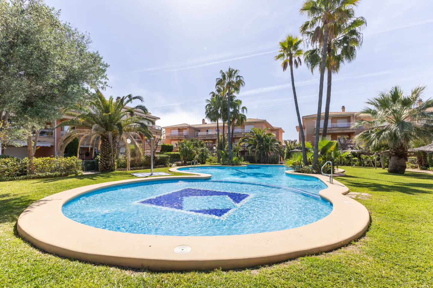Outdoor pool with blue mosaic design, surrounded by palm trees and green grass, with residential buildings in the background.